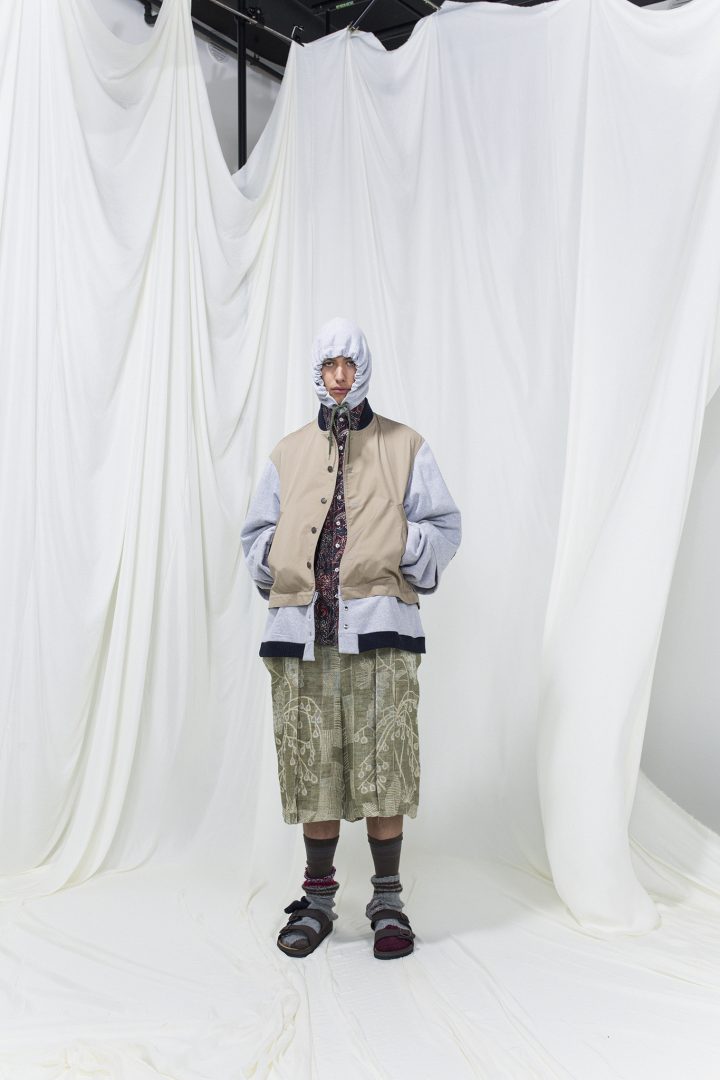 Model is wearing a panelled lilac and beige jacket with hoodie. Underneath green printed trousers and purple printed shirts. High socks and sandals as accessories