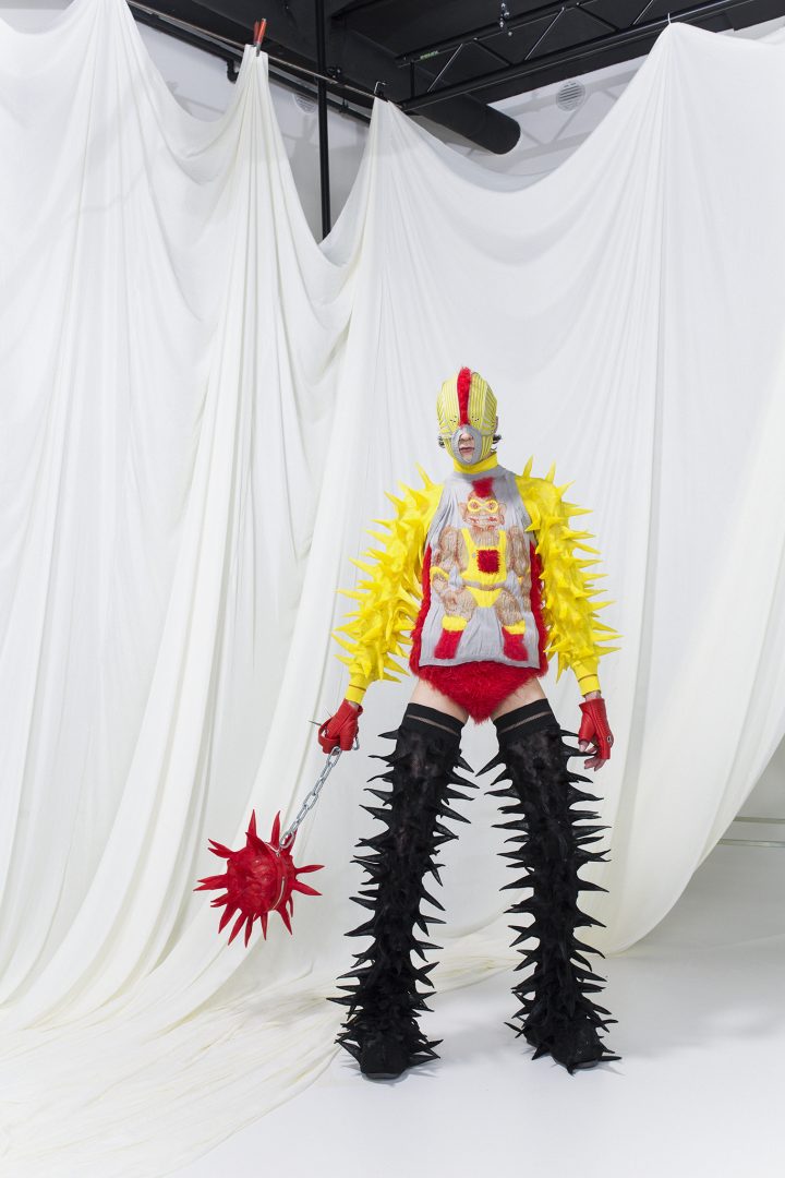 Model is wearing a mohair troll-doll tanktop with matching mask and red mohair briefs. Yellow spike bolero and black spike stay ups. Red leather gloves and spike bag.