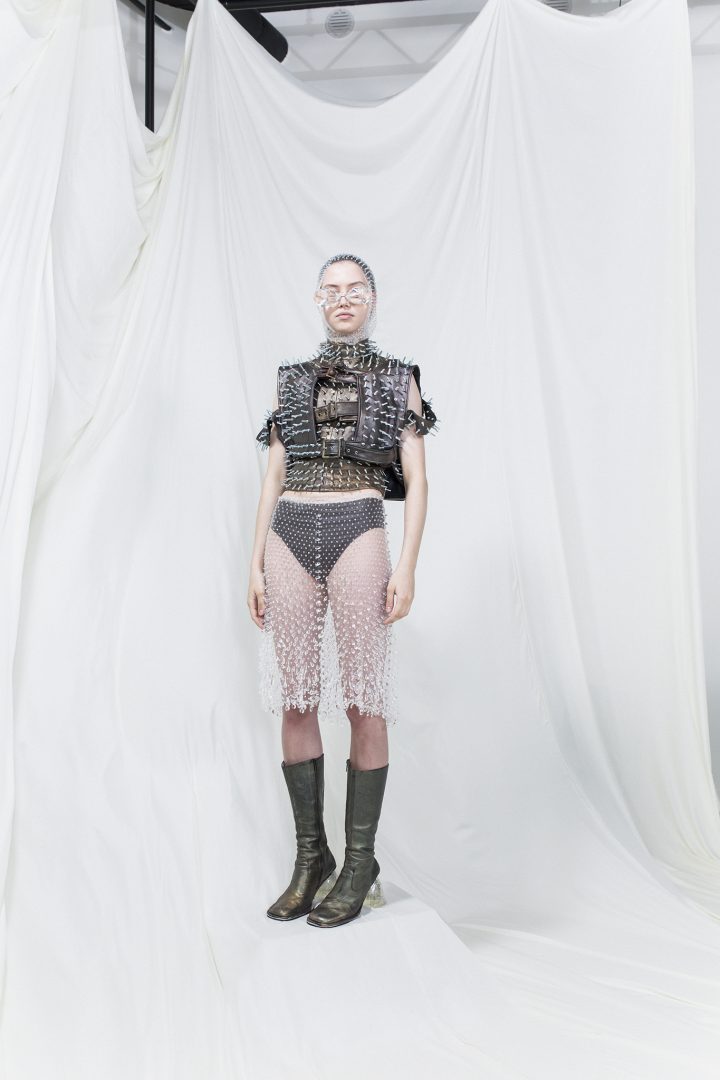 Model is wearing a spiked brown and green leather vest with buckles, sheer sequinned skirt and scarf