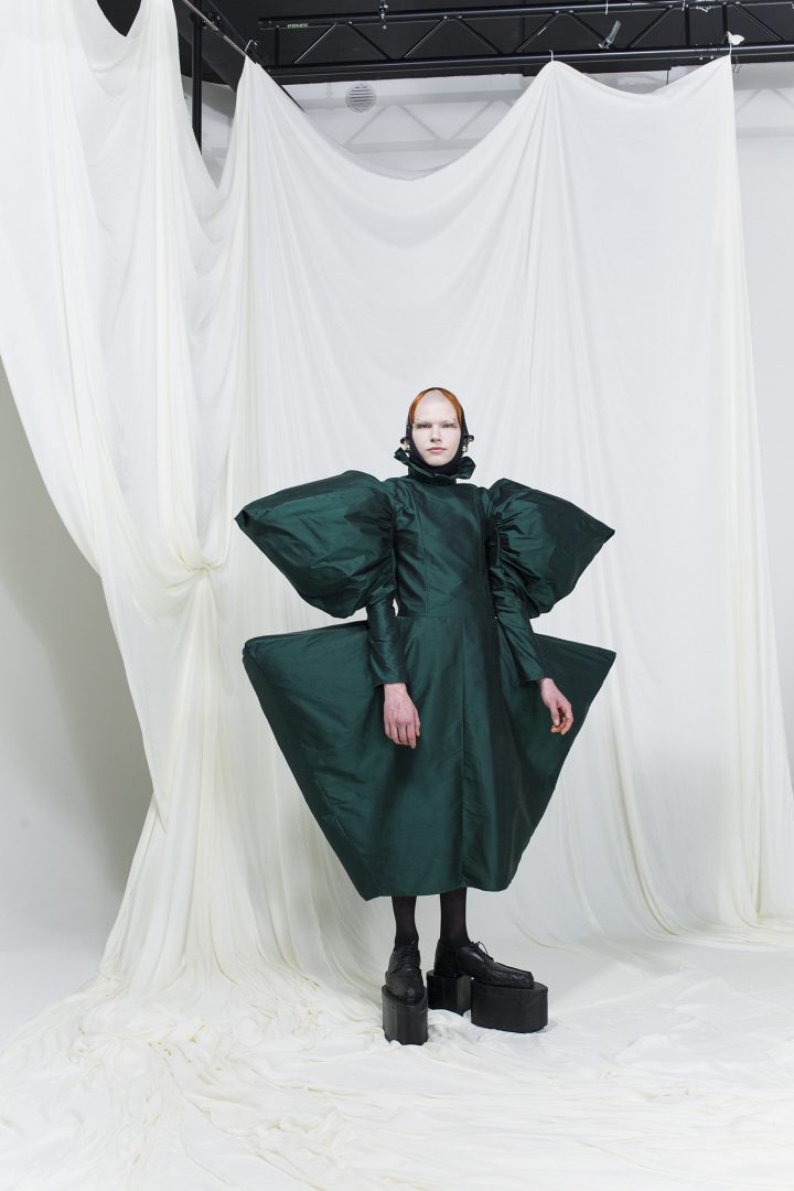 Model is wearing a emerald green dress with puffy angular shoulders and wide hips